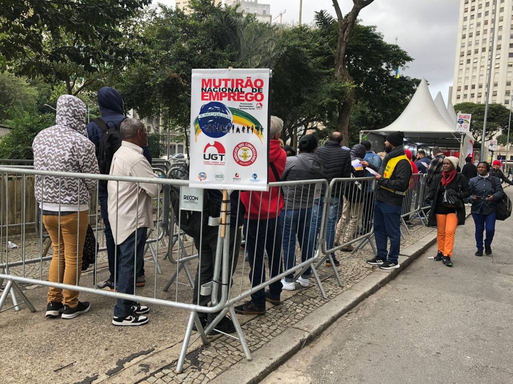 mutirão nacional de emprego A imagem mostra uma fila de pessoas ao ar livre, aguardando para participar de um mutirão nacional de emprego. As pessoas estão vestidas com roupas de frio, indicando que o clima é ameno ou frio. Algumas delas estão encostadas em grades de contenção, que organizam a fila. No centro da imagem, há um cartaz com a inscrição