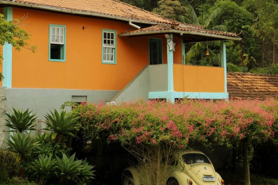 casa amarela na frente de uma floresta com várias arvores altas e verdes. embaixo da casa, um toldo rodeado por flores rosas claras, e um fusca amarelo estacionado embaixo.