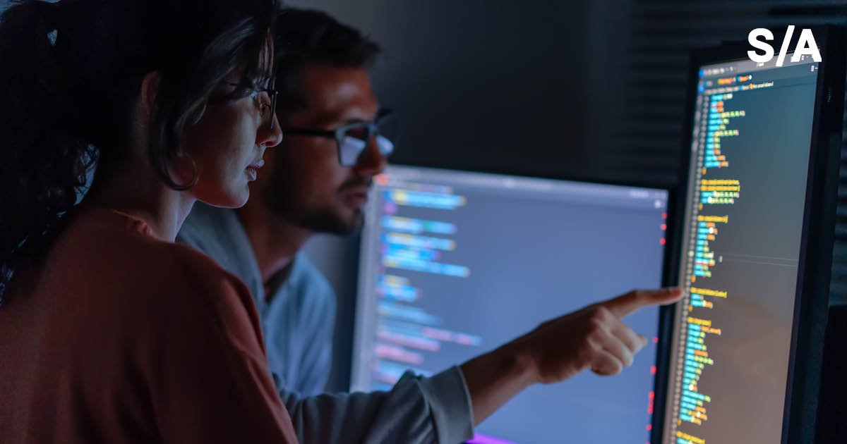 Dois colegas de trabalho conversam em frente a uma tela de computador com dados de programação.