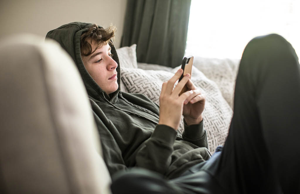 Foto de um adolescente mexendo no celular enquanto esta sentado no sofa.