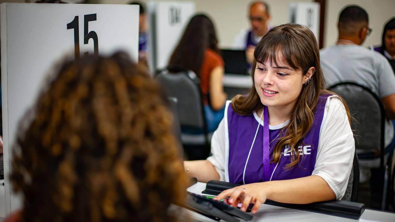 menina branca, com franja usando uma blusa branca e um colete azul escrito "CIEE" por cima olha para um teclado de computador e sorri.