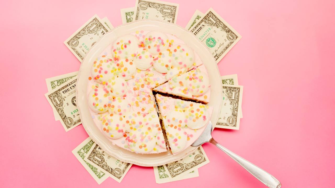 Visão superior de um bolo de aniversário, sendo fatiado em cima de notas de dólares.